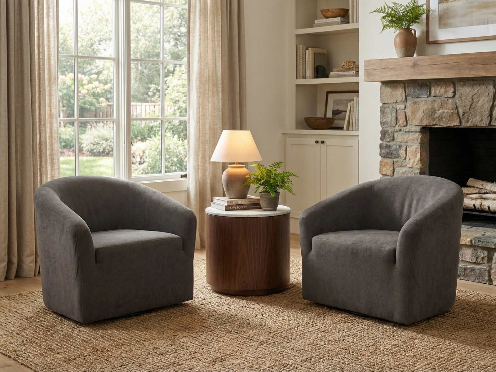 Two matching Bentley Easy Chairs, upholstered in dark gray fabric with a continuous, curved silhouette, are placed facing each other over a woven jute rug. They flank a modern cylindrical side table with a wood base and white marble top, which holds a lamp and a potted fern. The seating area is situated in front of a large window with sheer curtains and a rustic stone fireplace with a wooden mantelpiece, creating a cozy living room scene