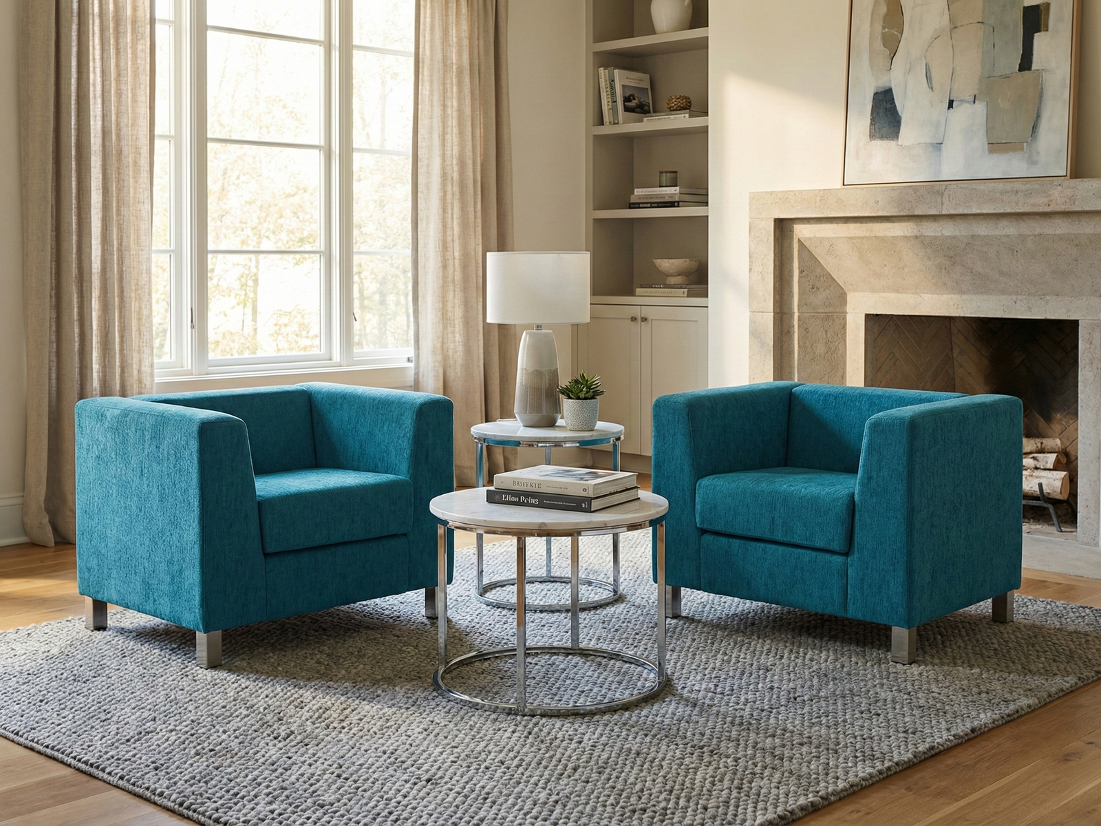 Two matching Kim Easy Chairs, upholstered in bright textured teal fabric with a modern, boxy silhouette and short metallic legs, are placed facing each other over a gray woven rug. They flank a pair of nested round side tables with marble tops and chrome frames, holding a lamp and stacked books. The setting is a brightly lit living room with a large window, neutral curtains, a built-in bookshelf, and a stone fireplace with contemporary artwork above it