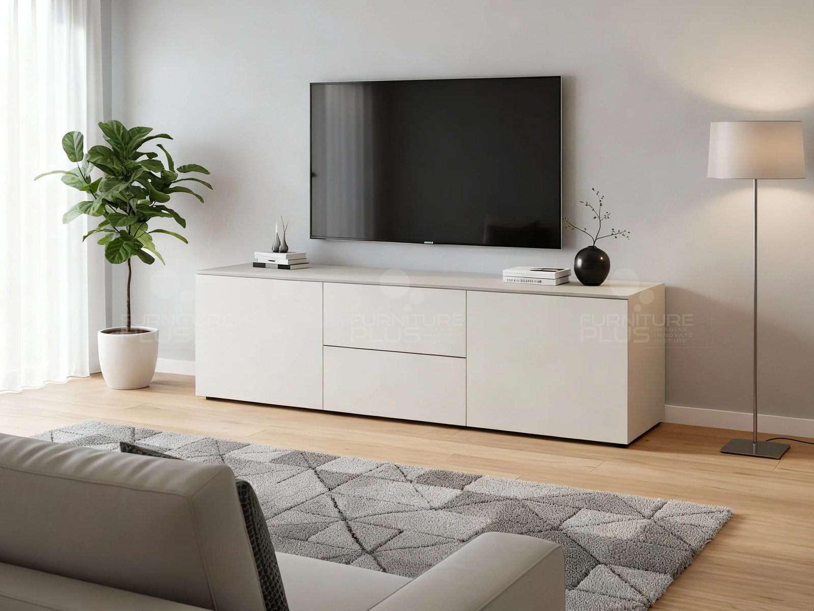 A minimalist living room featuring a long, low, off-white Nero TV console or sideboard with handleless cabinet doors and a central drawer, set against a light gray wall. A large flat-screen television is mounted above the unit. The console is styled with minimalist decor, flanked by a tall potted fiddle-leaf fig tree on the left and a modern floor lamp on the right. A plush gray and white geometric rug and the back of a light-colored sofa are visible in the foreground