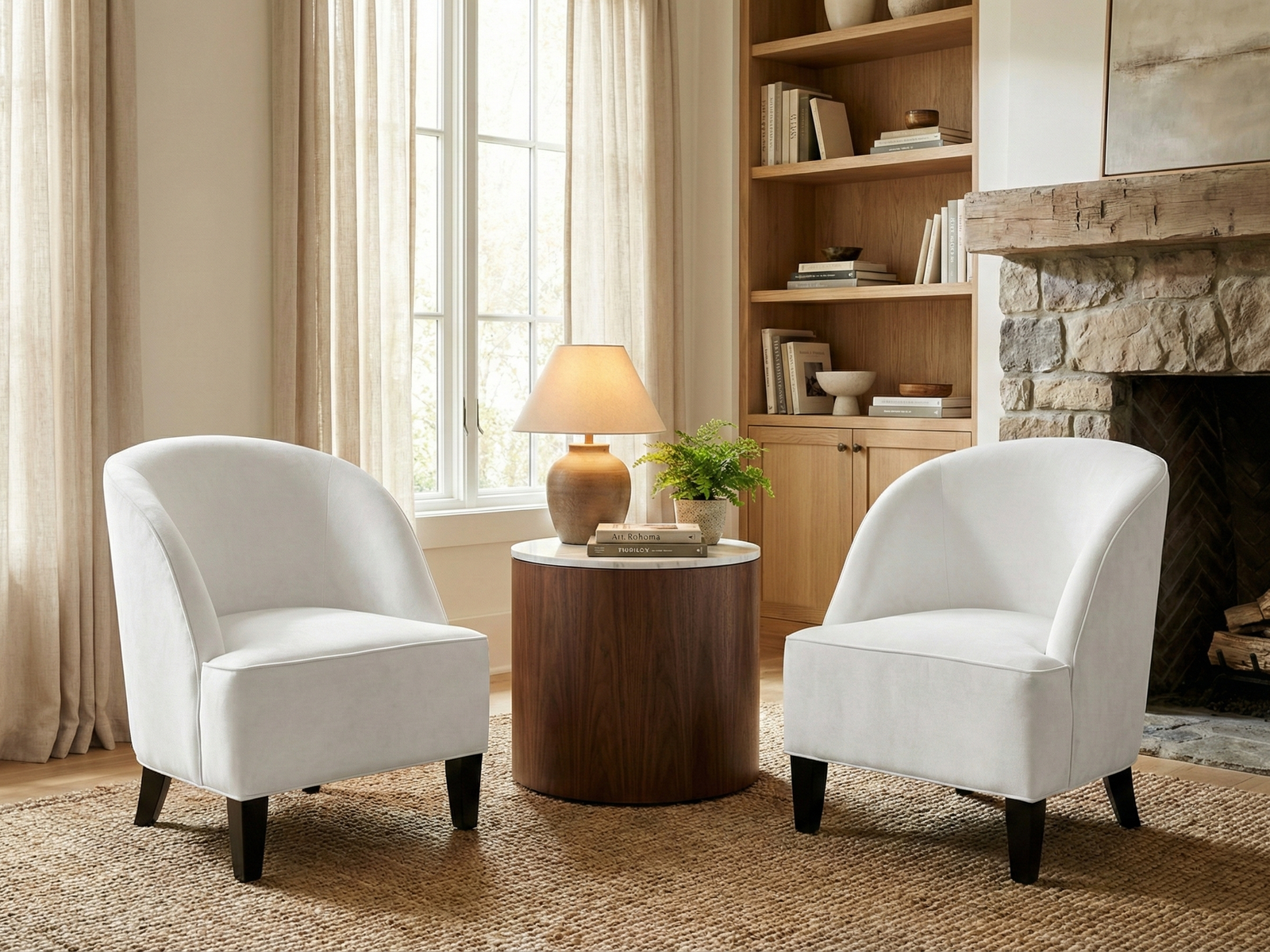 Two matching Smith Easy Chairs, contemporary tub armchairs upholstered in white fabric with curved backs and low arms, are positioned facing inward over a woven jute rug. They flank a modern cylindrical side table with a wood base and a white marble top, holding a warm-toned ceramic lamp and a small potted fern. The seating area is set in a cozy living room with a large window draped in sheer curtains, a built-in wooden bookshelf, and a rustic stone fireplace with a wood mante