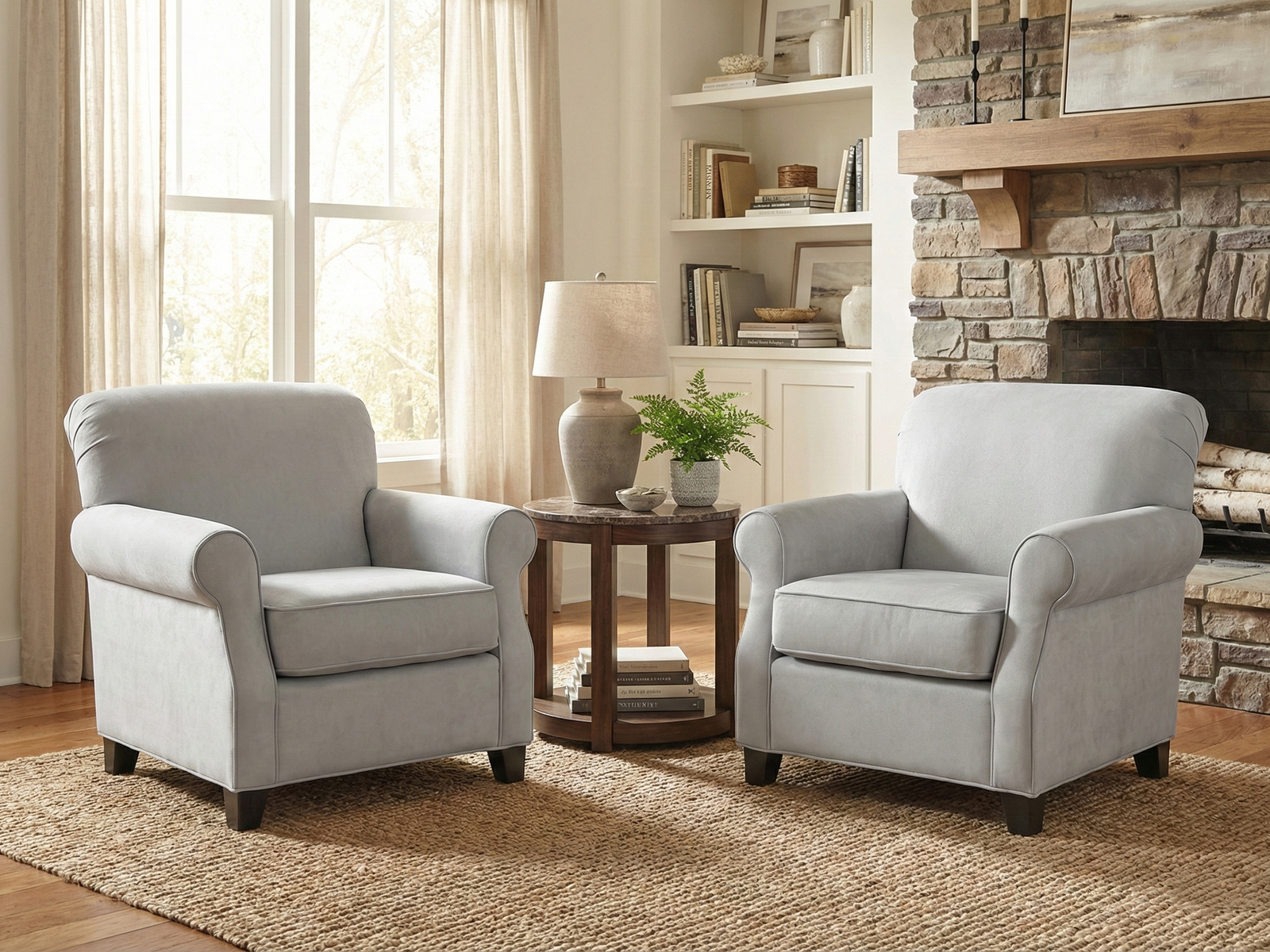 Two matching Stacy Easy Chairs, traditional armchairs upholstered in light gray fabric with rolled arms and backs, are placed facing each other over a woven jute rug. They flank a round wooden side table with a marble top, which holds a ceramic lamp and a potted fern. The chairs are set in a cozy living room with a large window draped in sheer curtains, a built-in bookshelf, and a rustic stone fireplace with a wooden mantel