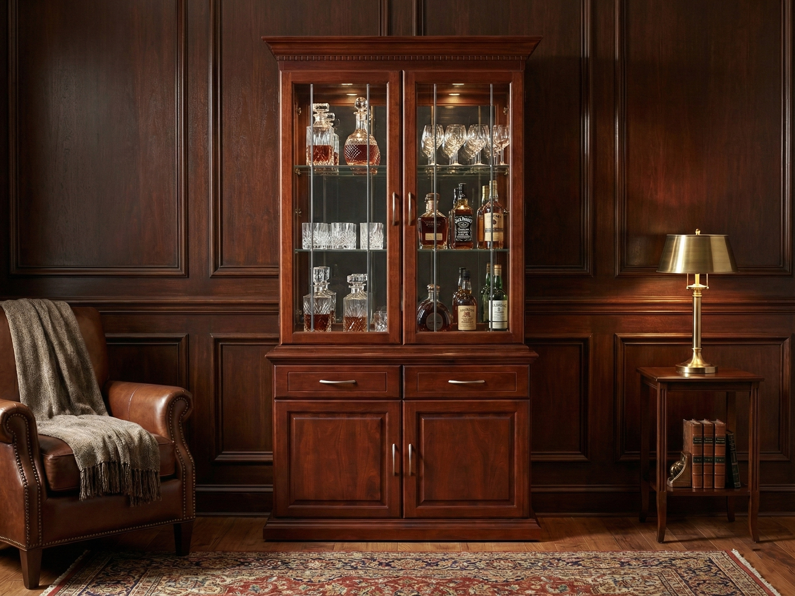 A cherry-toned wooden Kentone 2 Glass liquor hutch with crown molding. The upper section displays barware behind glass doors with vertical mullions, staged in a classic study with a leather club chair and a brass table lamp