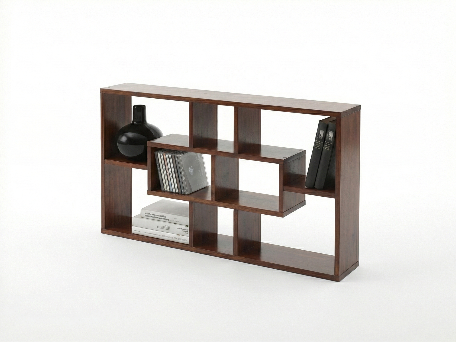 Side view of a low, wide, dark wood Upney display shelf unit with a modern, open-frame design. The internal shelving forms a nested, asymmetric, rectangular pattern. The unit is styled with a large black vase, a stack of CDs, and several books, set against a plain white background