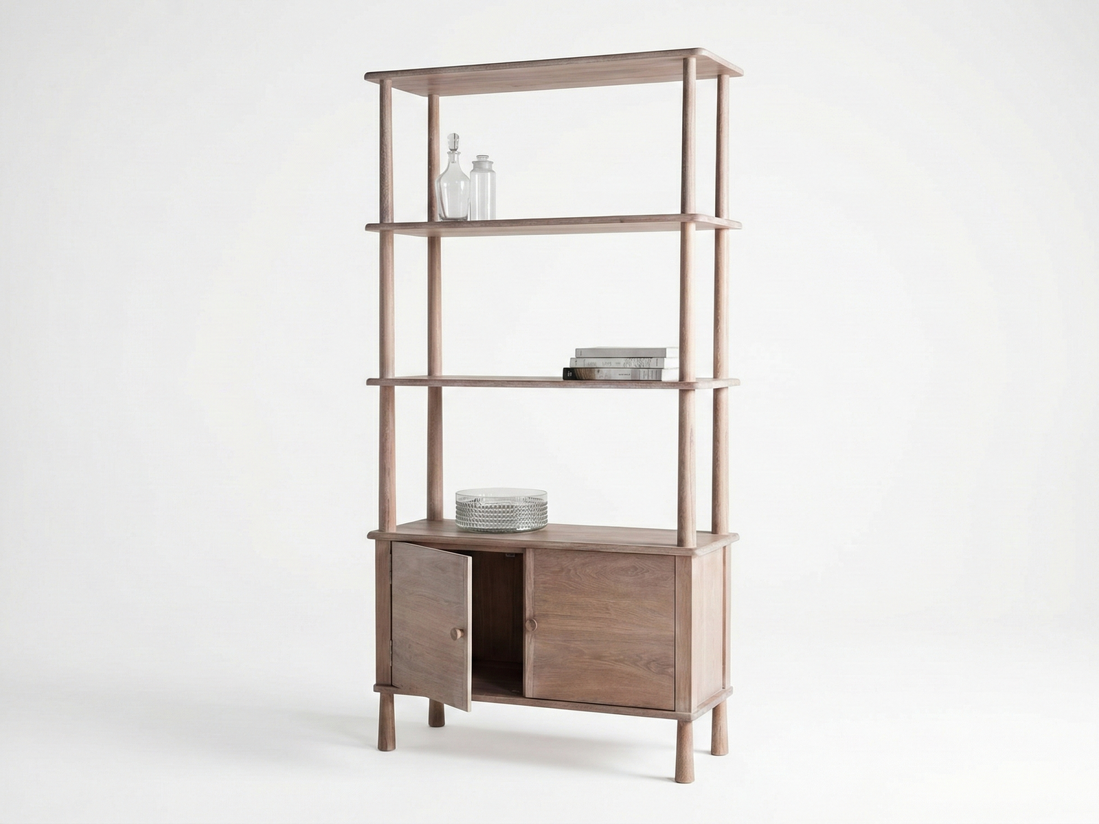 Side view of a tall, light-brown wooden display unit (Woodford style) with a modern, open-shelving design over a two-door cabinet base. The unit features four open shelves supported by rounded vertical poles and two cabinet doors at the bottom, one of which is slightly ajar. The shelves are minimally styled with two clear glass bottles, a stack of books, and a stack of clear plates, all set against a white background