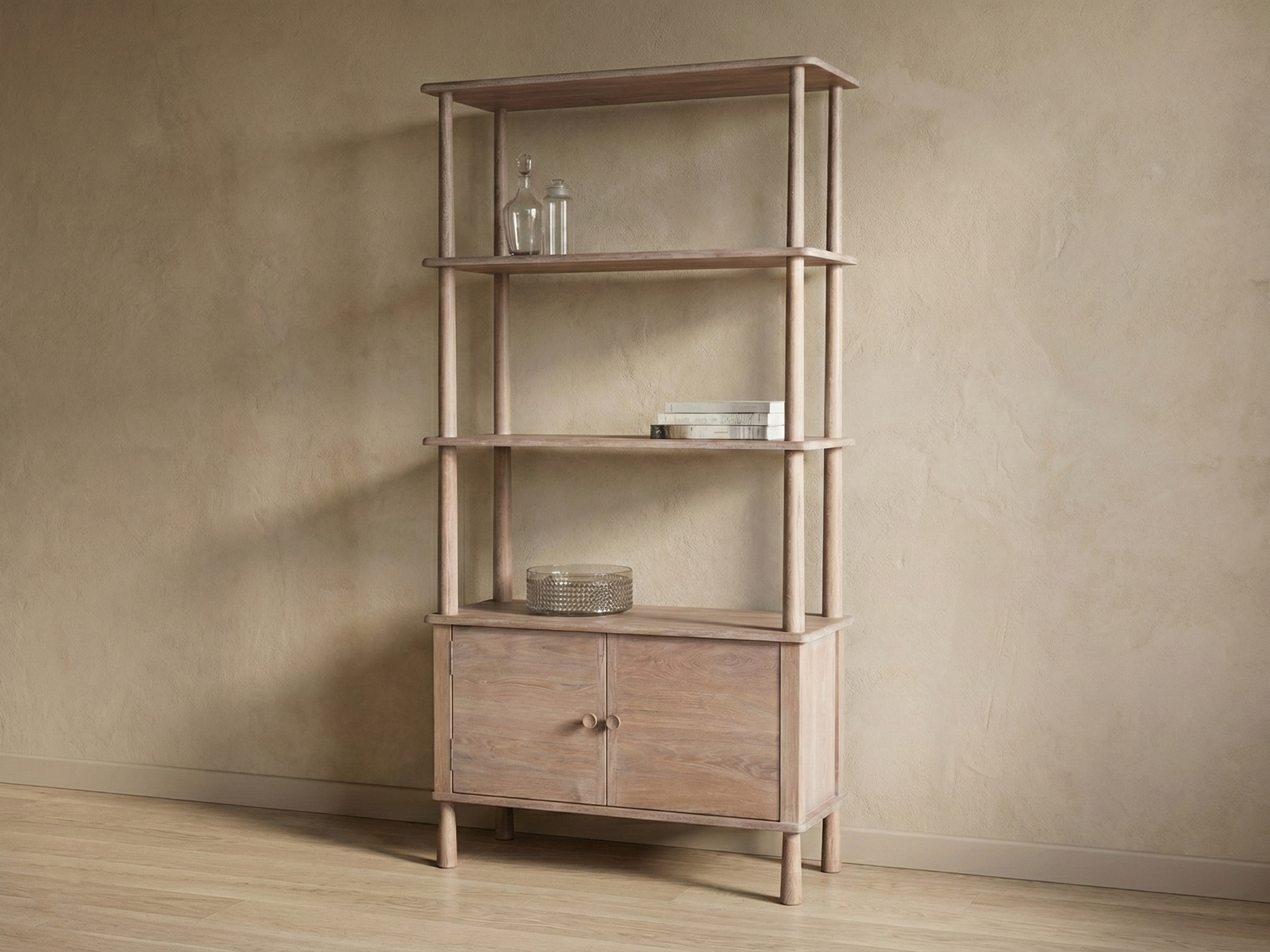A tall, light-brown wooden display unit (Woodford style) with a modern design, set against a warm beige textured wall and a wooden floor. The unit features four open shelves supported by rounded vertical poles and a two-door cabinet base with rounded legs. The shelves are minimally styled with two clear glass bottles, a stack of books, and a textured clear glass bowl on top of the cabinet section